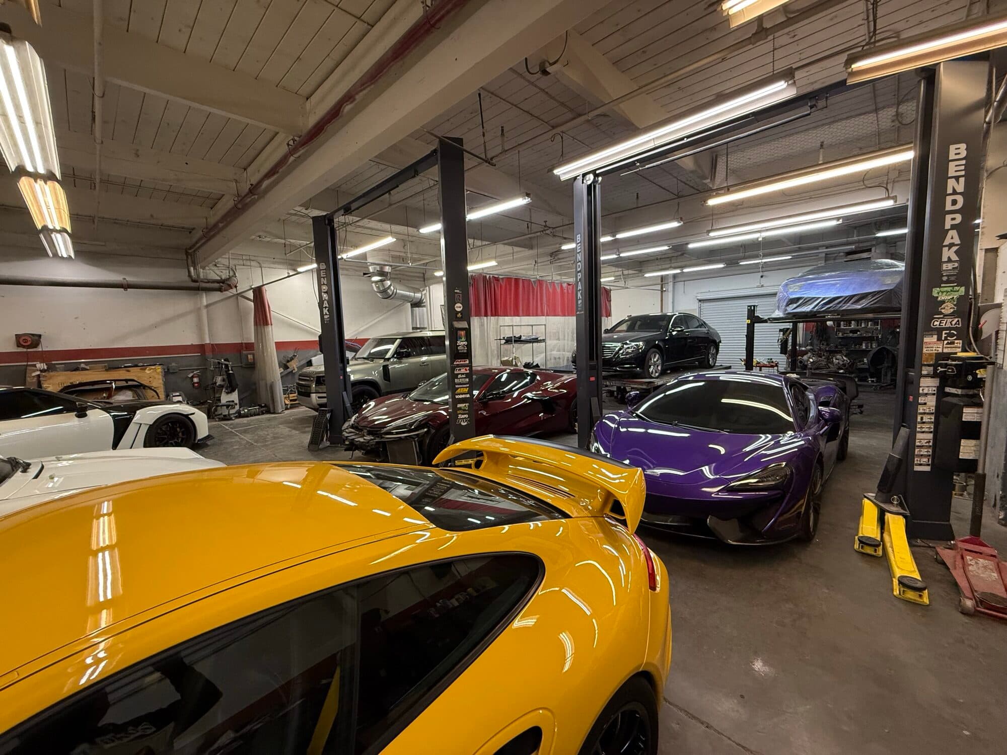 A yellow Porsche 911 GT2 with two McLarens up on the lifts behind it at Alpina Auto Body, exotic collision and body shop in Southeast Portland, Oregon
